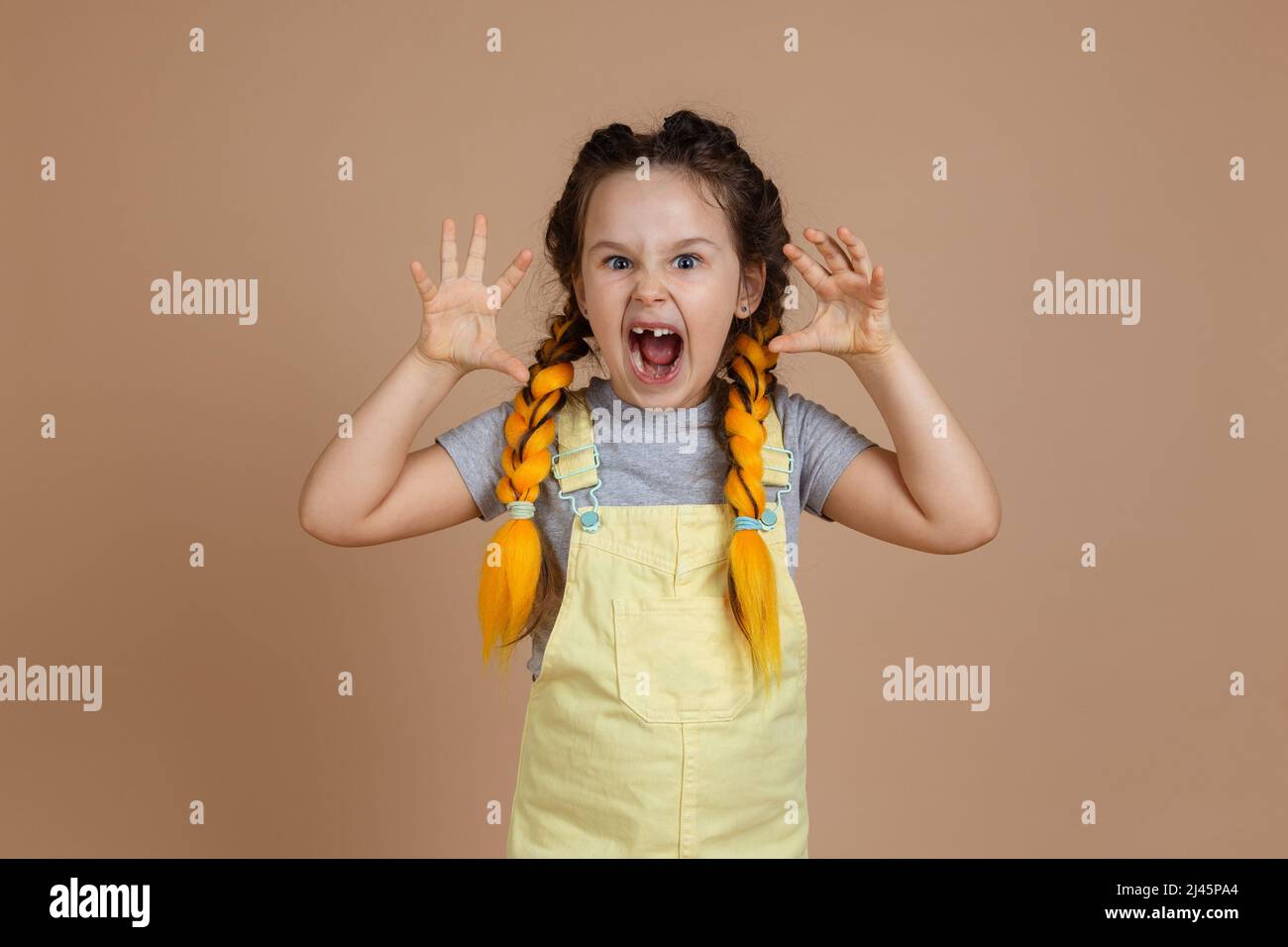 Porträt eines verspielten kleinen Mädchens mit gelben Kanekalon-Zöpfen, das sich mit Händen und geöffnetem Mund in gelbem Jumpsuit als furchterregend einstellt Stockfoto