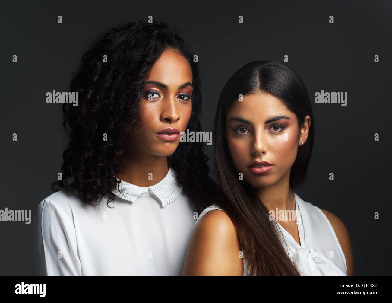 Es gibt Schönheit in einem Frauenfokus. Portrait von zwei attraktiven jungen Frauen in weißen Blusen, die eng zusammen vor einem dunklen Hintergrund in posieren Stockfoto