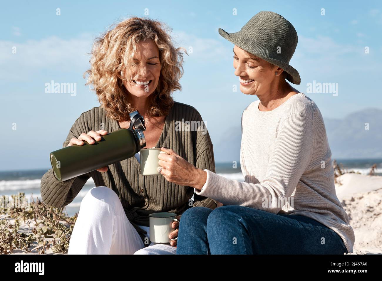 Kann ich Euch auftoben? Eine kurze Aufnahme von zwei attraktiven reifen Frauen, die am Strand sitzen und Kaffee genießen. Stockfoto