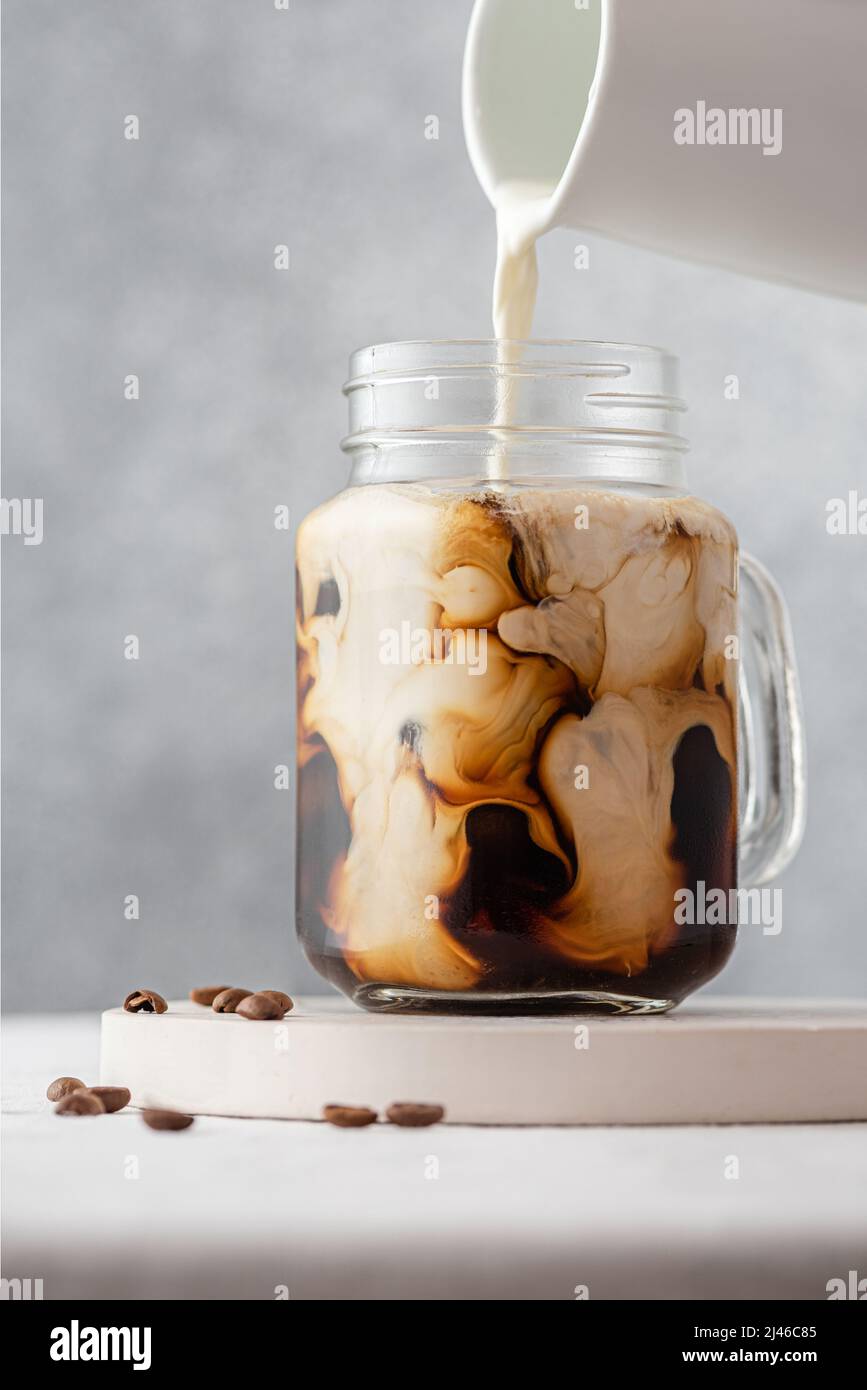 Gieße Milch oder Rahm in einem Glas auf grauem Hintergrund in den Eiskaffee. Kalte Erfrischung Sommergetränk. Vertikale Ausrichtung Stockfoto
