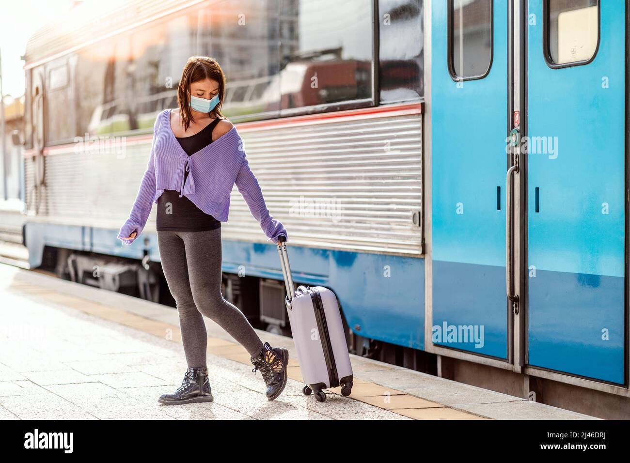 Eine junge Frau in einer Maske und mit Gepäck, die auf dem Bahnsteig eines Bahnhofs an ihrem Bestimmungsort läuft Stockfoto