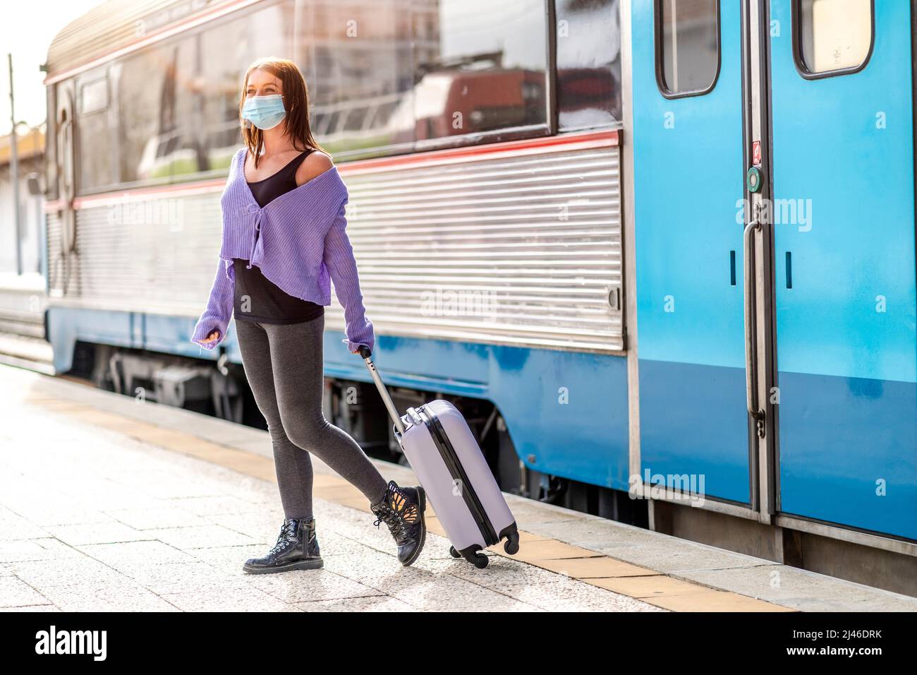 Eine junge Frau in einer Maske und mit Gepäck, die auf dem Bahnsteig eines Bahnhofs an ihrem Bestimmungsort läuft Stockfoto
