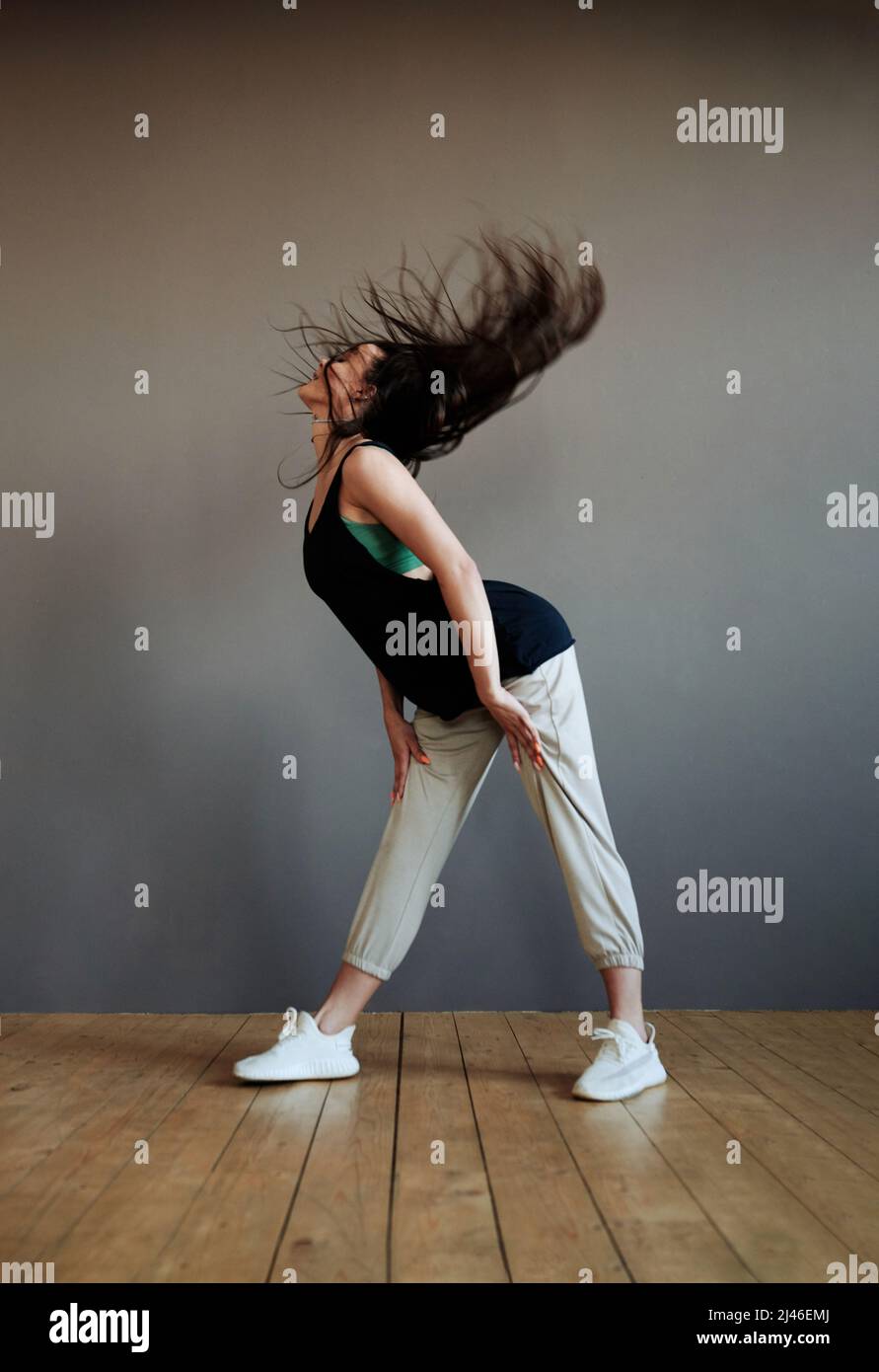 Zeitgenössische weibliche Teenager-Performerin in aktivem Ohr übt modische Tanzübungen, während sie im Studio gegen die graue Wand trainiert Stockfoto