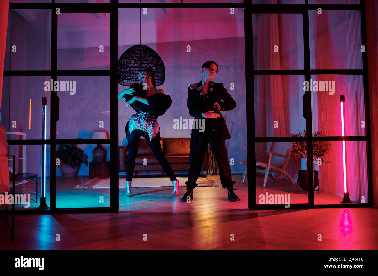 Zwei dynamische Teenager in schickem Gewand, die im Loft-Studio, in der Wohnung oder im Nachtclub mit pinken und blauen Neonlichtern modischen Tanz aufführen Stockfoto