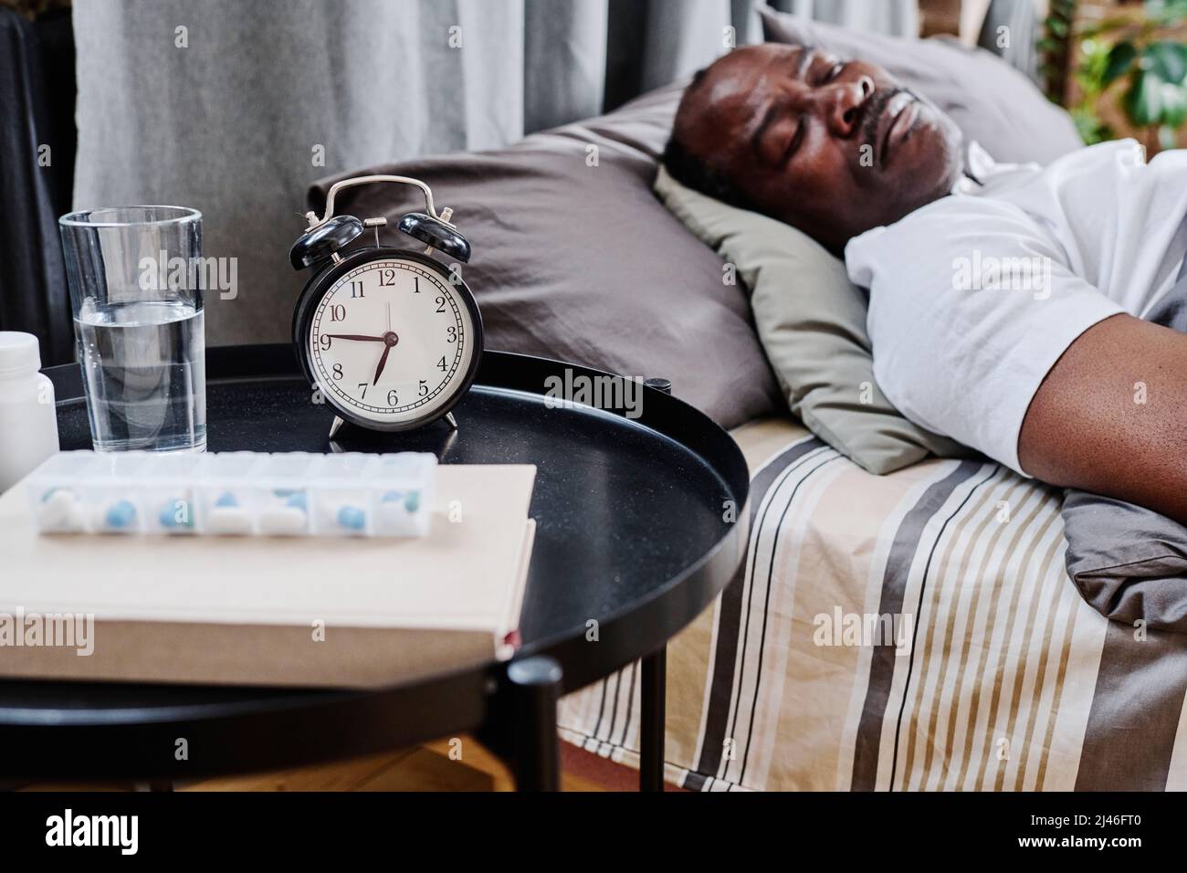 Kleiner runder schwarzer Tisch mit Wecker, Medikamenten, Buch und einem Glas Wasser, der neben dem Doppelbett steht, wo ein älterer Mann schläft Stockfoto