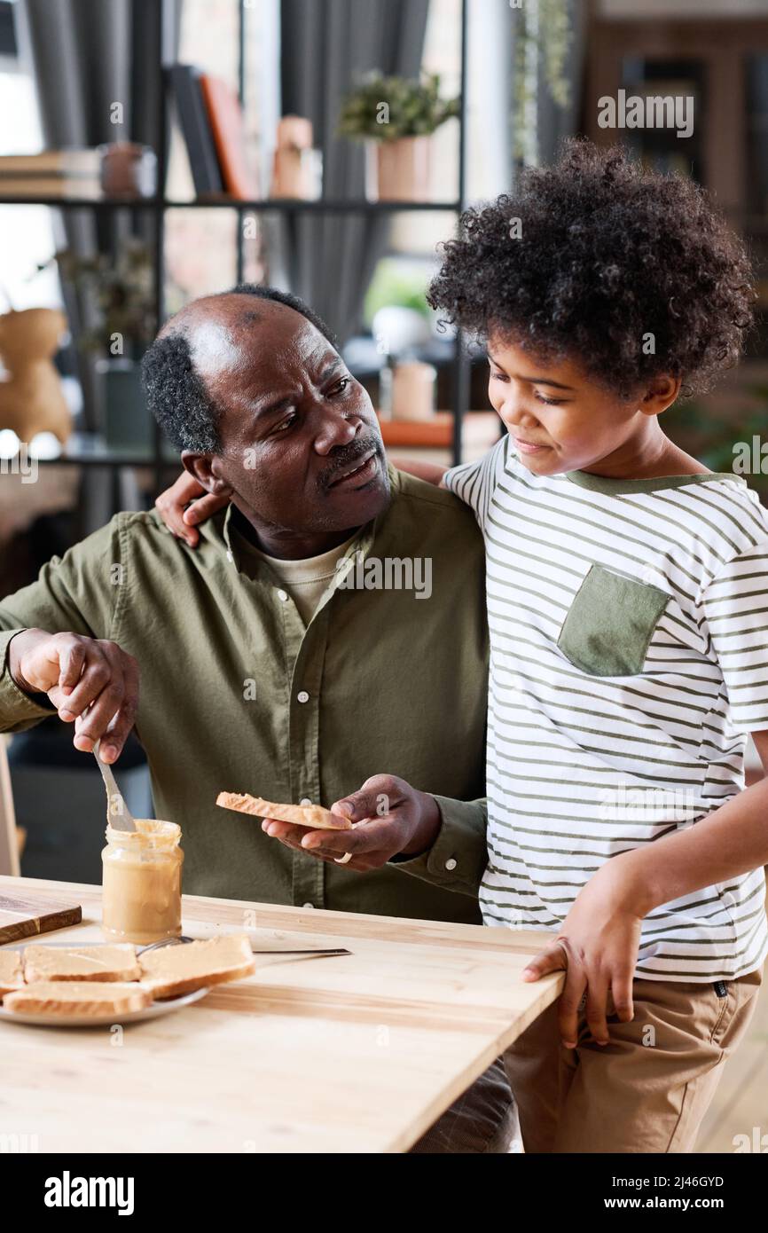 Netter multiethnischer Schuljunge, der bei seinem Großvater steht und ihm ein Sandwich mit Erdnussbutter zum Frühstück am Holztisch macht Stockfoto