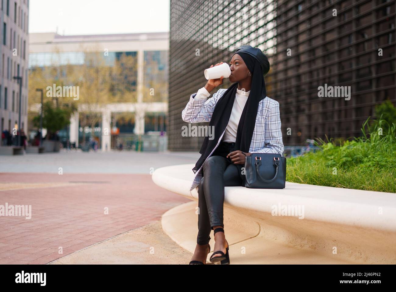 Stilvolle junge afroamerikanische Frau in elegantem legerem Outfit und schwarzem muslimischen Kopftuch, das auf einer Steinbank sitzt und Kaffee aus der Tasse im modernen Business Center trinkt Stockfoto