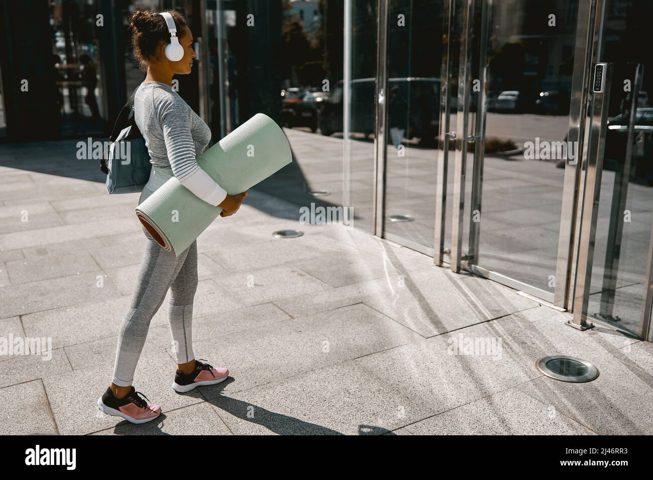 Sportliche Yoga-Frau in aktiver Kleidung und Kopfhörer beim Gehen mit Matte für das Training Stockfoto