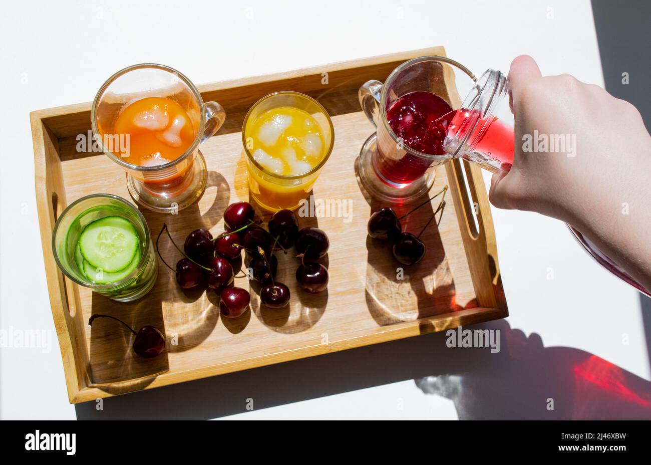 Ein Holztablett mit bunten Getränken: Orange, Gelb und Gurkenlimonade. In der Nähe sind Kirschen. Eine Hand gießt roten Saft aus einem Dekanter in ein Glas. C Stockfoto