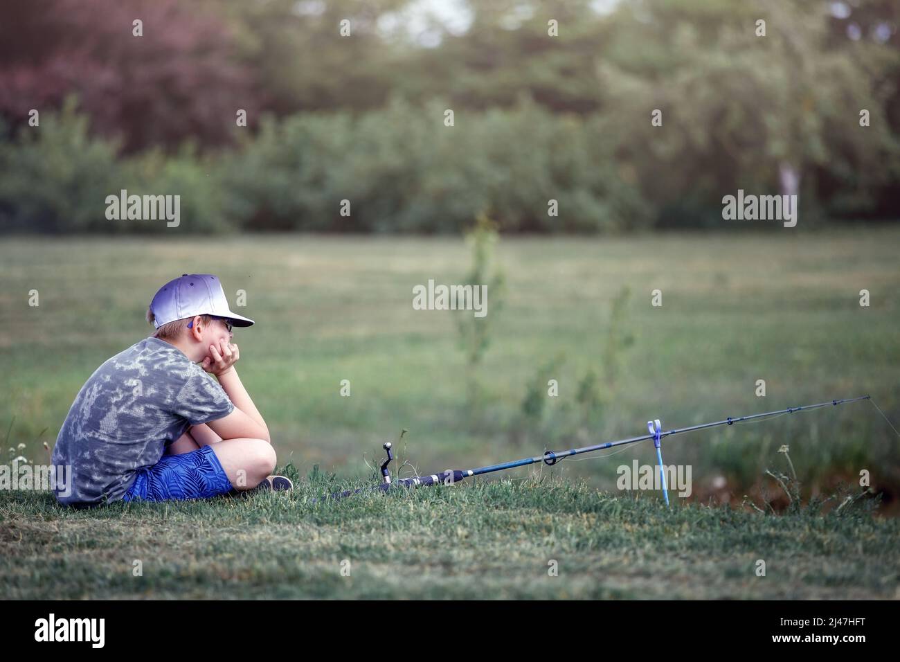 Naturforscher Junge sitzt in der Nähe des Teiches und wartet darauf, einen Fisch zu fangen. Geduldiger junger Fischer, der im Sommer die Natur genießt Stockfoto