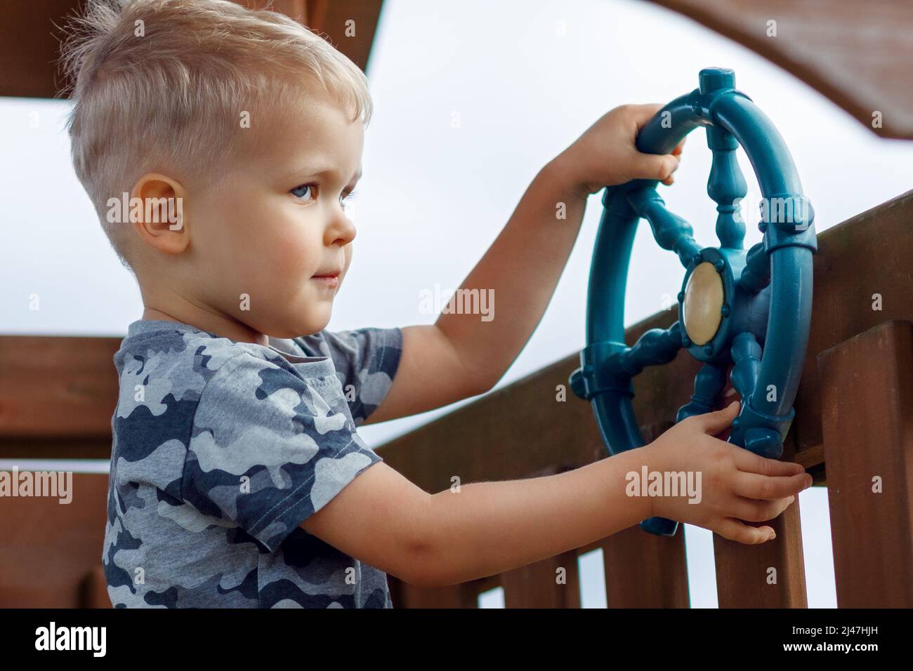 Freche Kinder fahren auf einem Spielplatz mit einem Schiffsrad. Das Kind in Camouflage fühlt sich in einem Holzspielhaus cool an. Kindheit voller Abenteuer Stockfoto