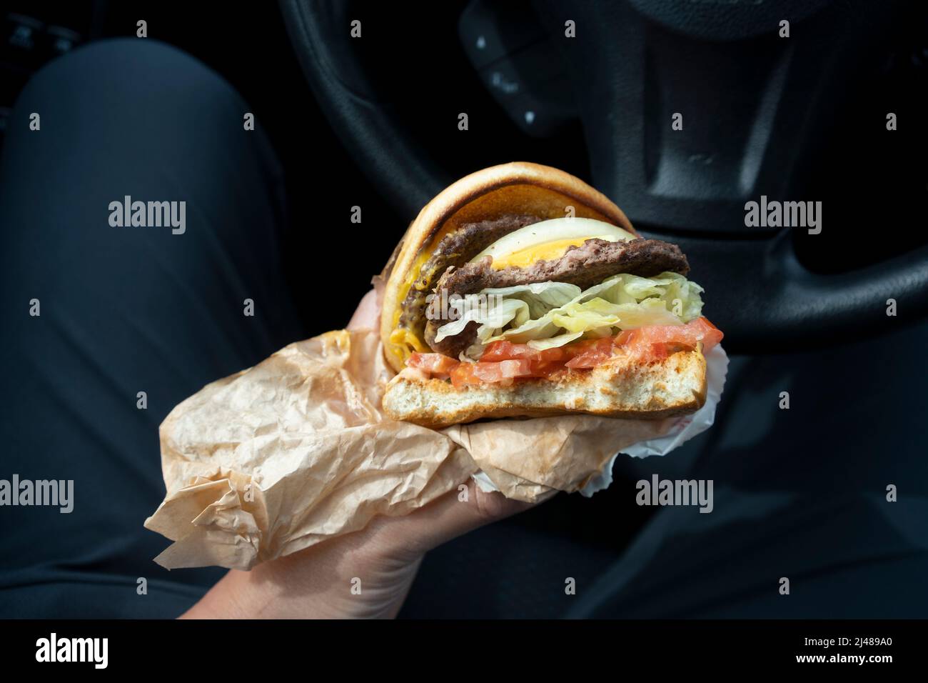 Eine Person isst in seinem Auto einen Cheeseburger mit doppeltem Patty, der von einem Drive-Through-Burger-Kettenrestaurant bestellt wurde. Gefährliche Chemikalien, bekannt als PFAS... Stockfoto