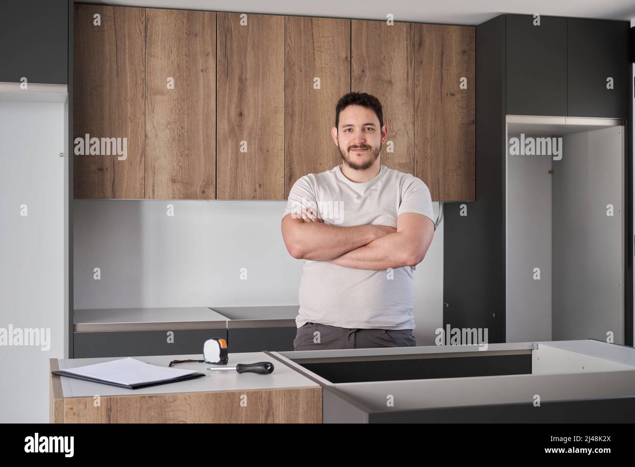 Ein junger hispanischer Mann lächelt und kreuzt seine Arme, nachdem er Küchenmöbel installiert hat. Stockfoto