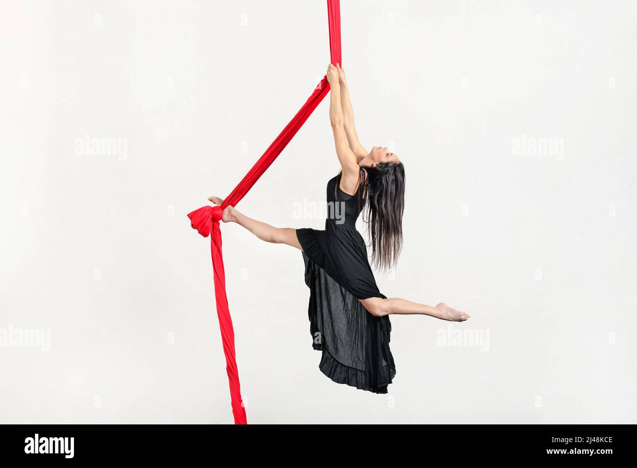 Ganzkörper-Seitenansicht einer aktiven weiblichen Luftakrobistin im Kleid, die im Studio auf roten Seiden vor weißem Hintergrund trickst Stockfoto