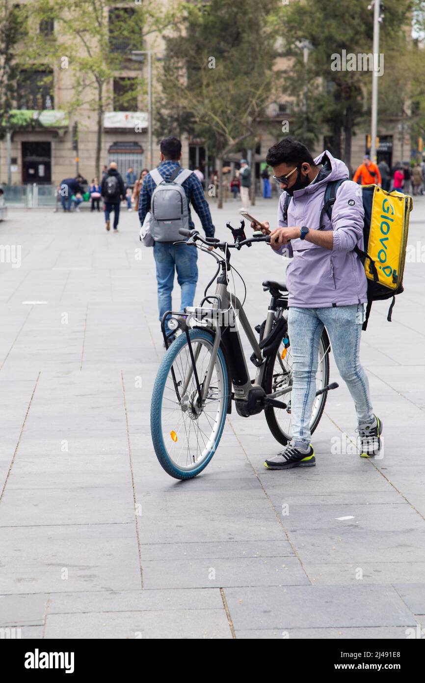 Männlicher Kurier in einer medizinischen Maske, um Nahrung zu liefern. Deliveryman mit Smartphone.Barcelona, Spanien - 09. April 2022 Stockfoto
