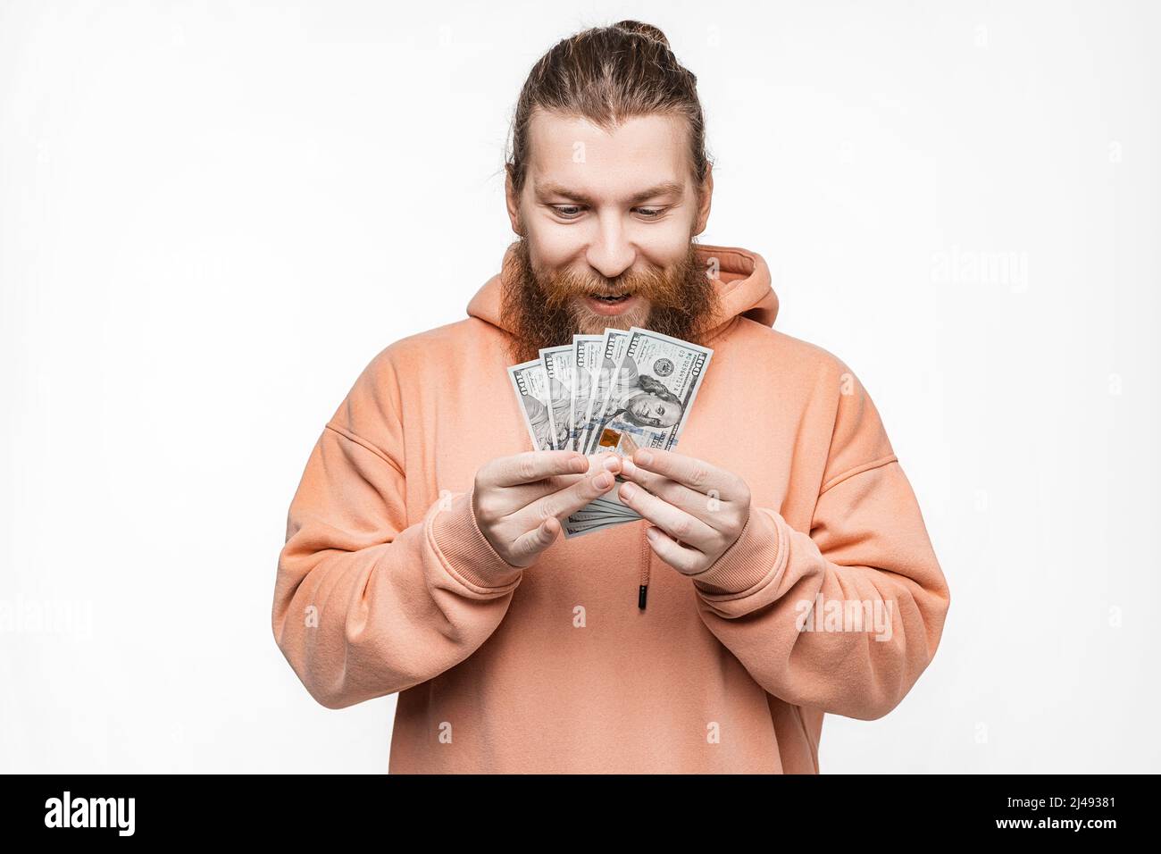 Hübscher skandinavischer Mann, der auf grauem Hintergrund Dollargeld in den Händen hält. Kerl mit Ingwer-Frisur und Bart in einem Sweatshirt. Die Konz Stockfoto