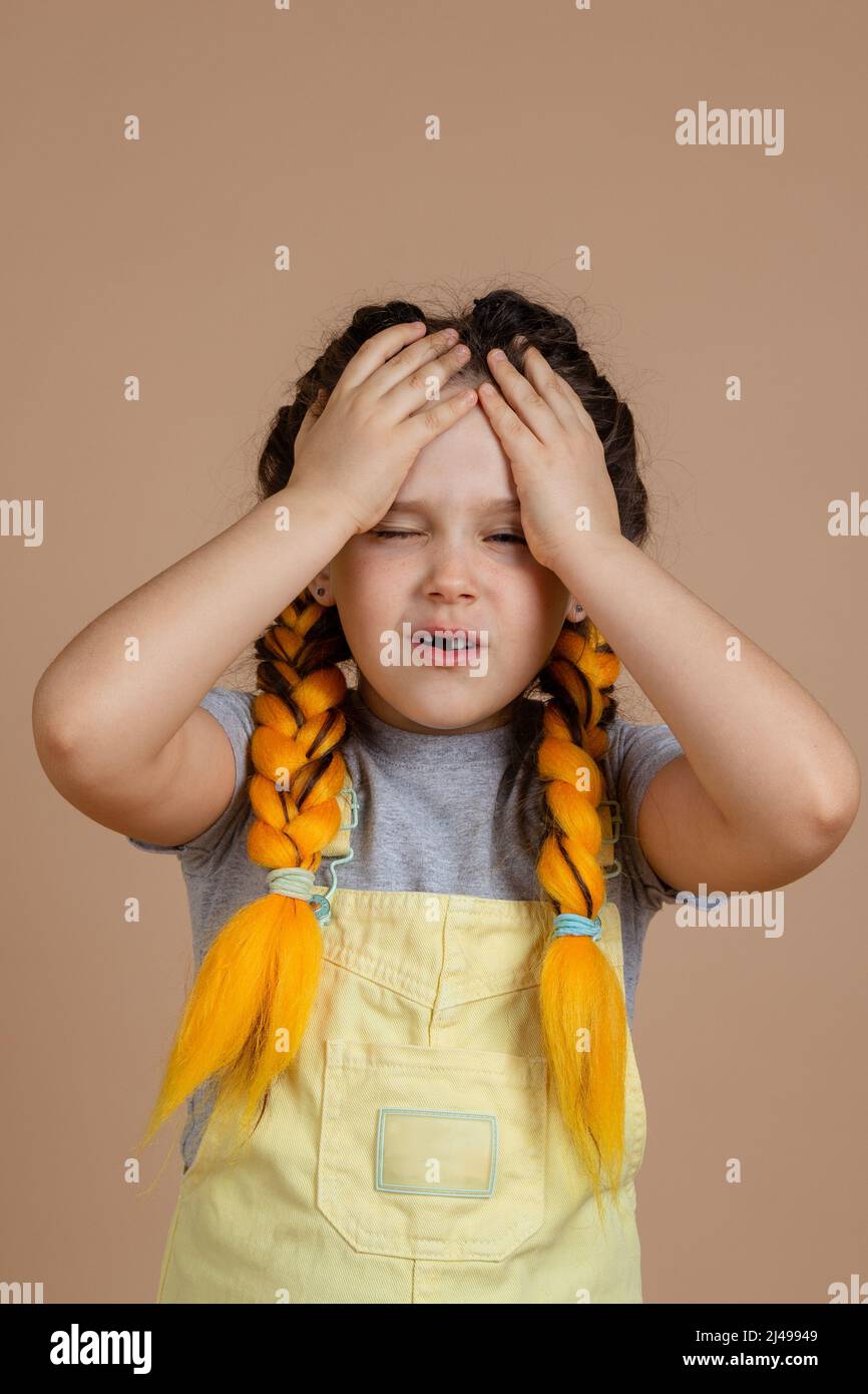 Kleines weibliches Kind mit gelben Kanekalon-Zöpfen, Kopfschmerzen, die den Kopf mit den Händen berühren und mit halbgeöffnetem Mund und Augen in gelbem Jumpsuit und Stockfoto