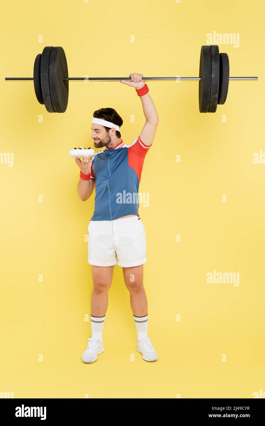 Seitenansicht des jungen Sportlers, der Kuchen riecht und die Hantel auf gelbem Hintergrund hebt Stockfoto