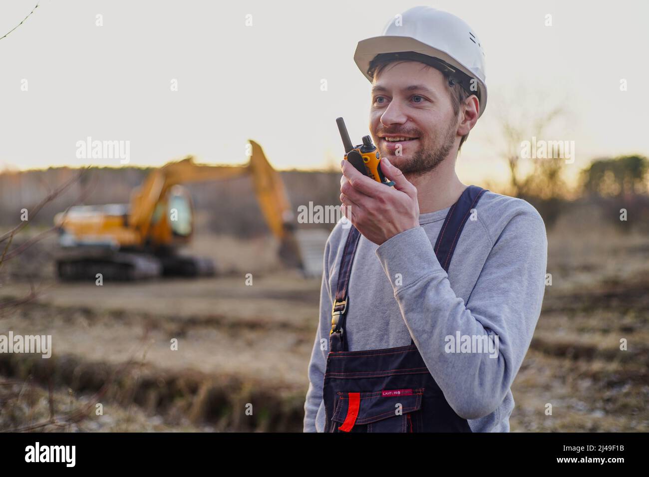 Lächelnder Baumeister im Helm, der durch den Funksender auf der Baustelle spricht. Bagger im Hintergrund. Stockfoto