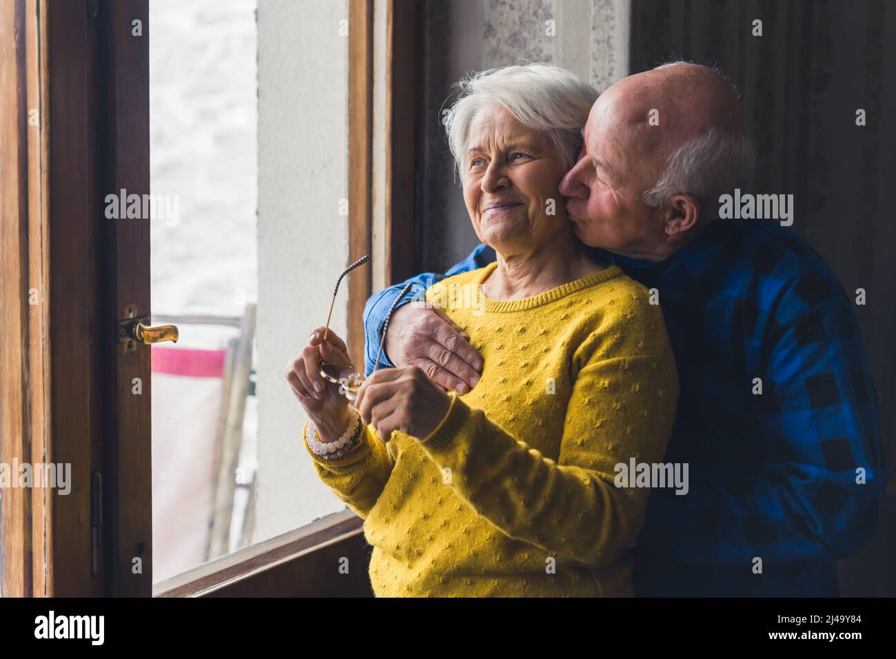 Älteres kaukasisches Paar am Fenster. Glückliche Beziehung. Der alte kahle Mann umarmte seine Frau und gab ihr einen Kuss auf ihr Küken. Hochwertige Fotos Stockfoto