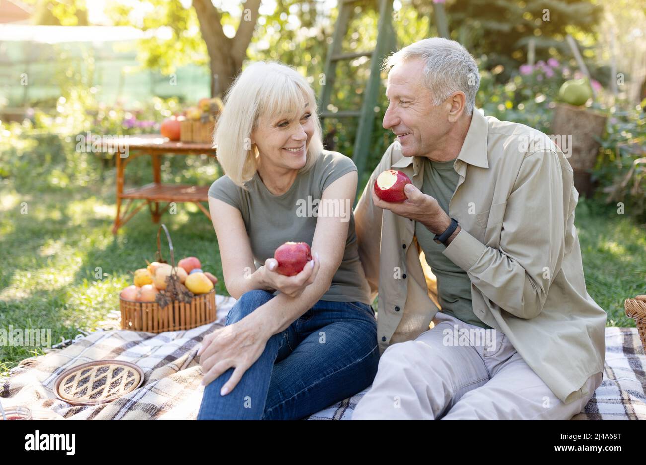 Gesunder und glücklicher Ruhestand. Positive ältere Ehepartner sitzen auf dem Rasen und essen Äpfel und ruhen sich im Garten aus Stockfoto