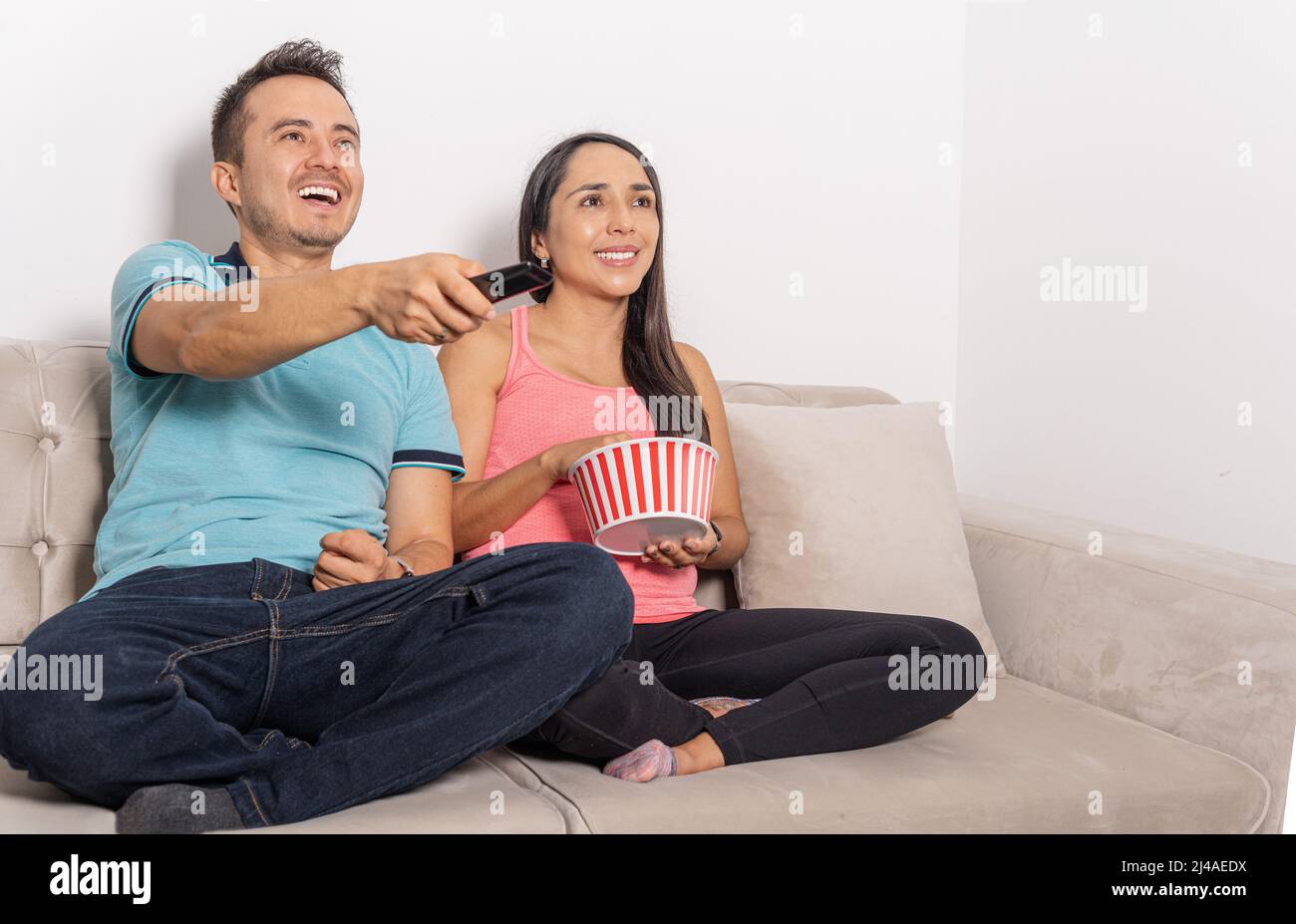 Latino-Mann und -Frau, die mit der Fernbedienung Fernsehen und Popcorn essen, die zu Hause auf einer Couch sitzen Stockfoto