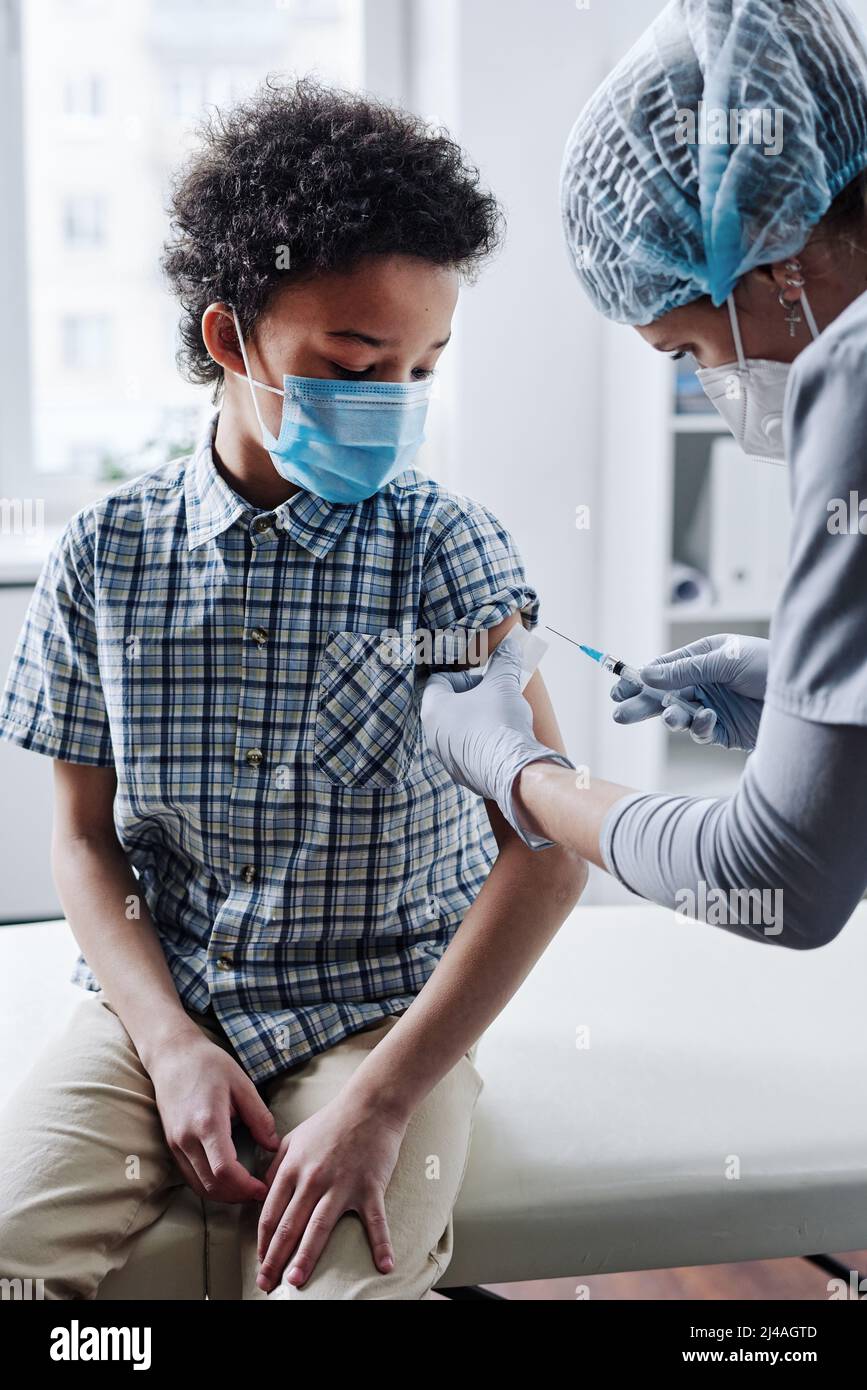 Das maskiert Kind erhält eine Impfung von der Krankenschwester, während es das Kinderbüro besucht Stockfoto