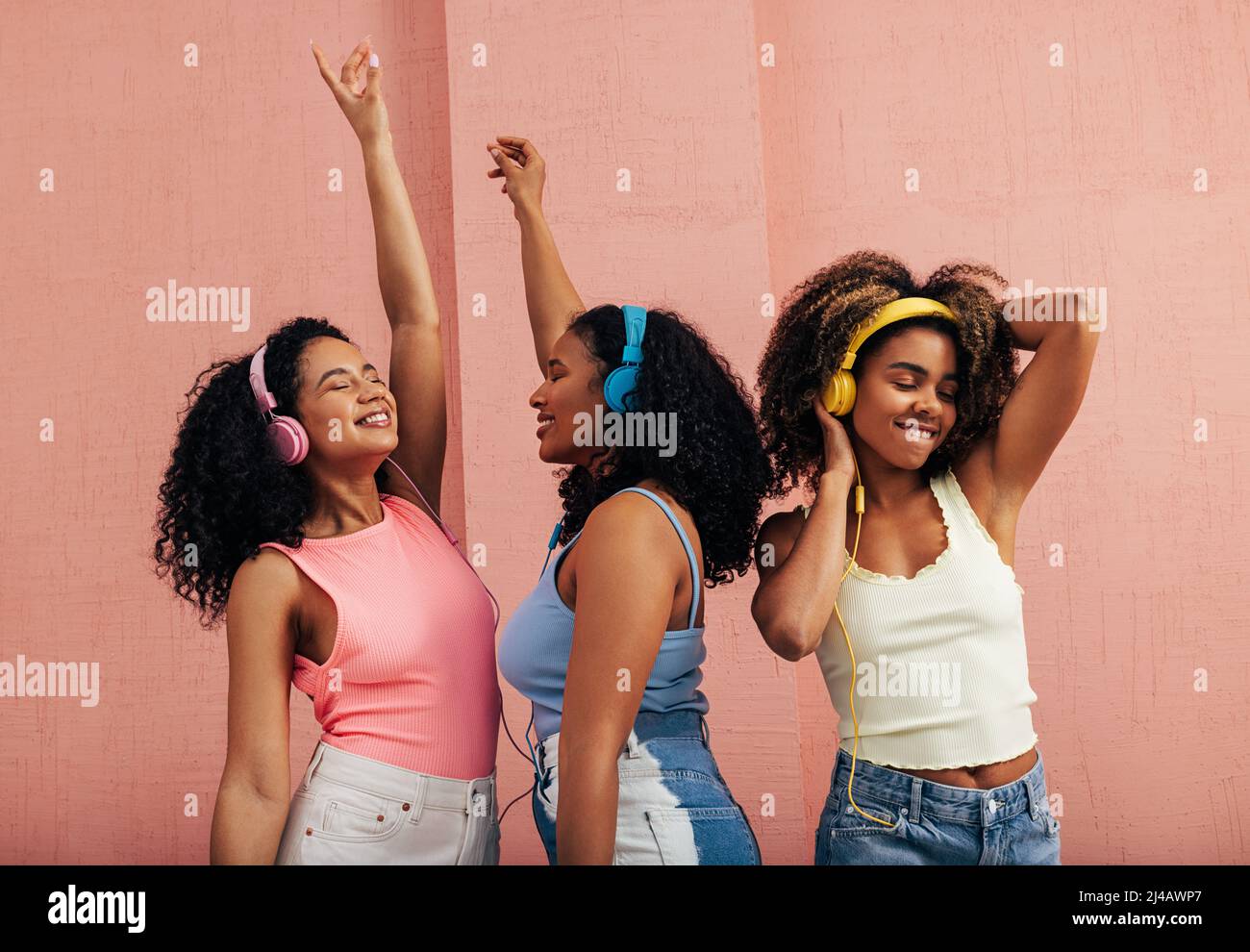 Drei Freundinnen mit verschiedenen Körpertypen tanzen zusammen. Lächelnde Frauen mit Kopfhörern, die Musik an der rosa Wand genießen. Stockfoto