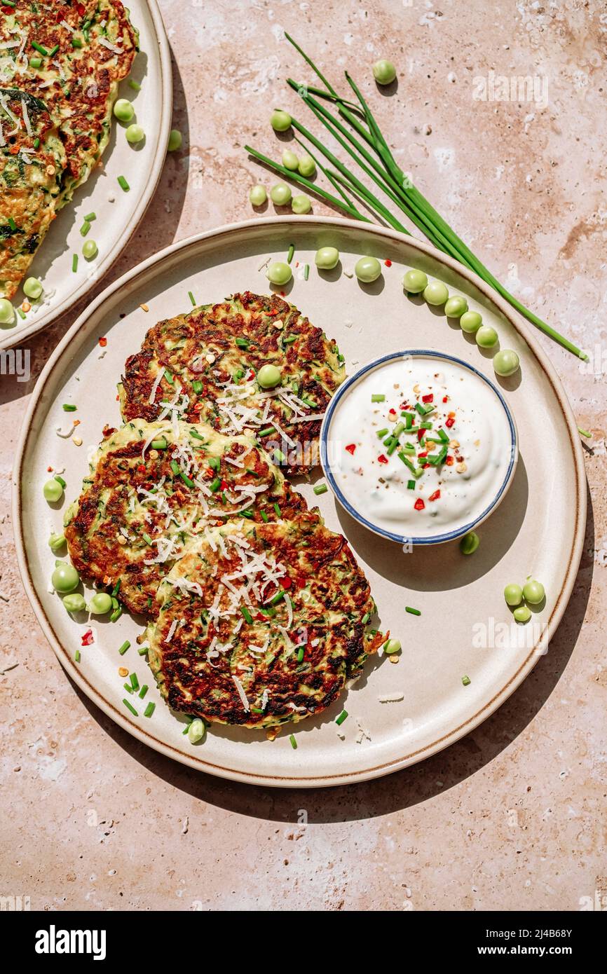 Zucchini-Pfannkuchen, Erbsen, Schnittlauch, geriebener Parmesan, Topf mit würziger Creme, auf weißem Teller Stockfoto