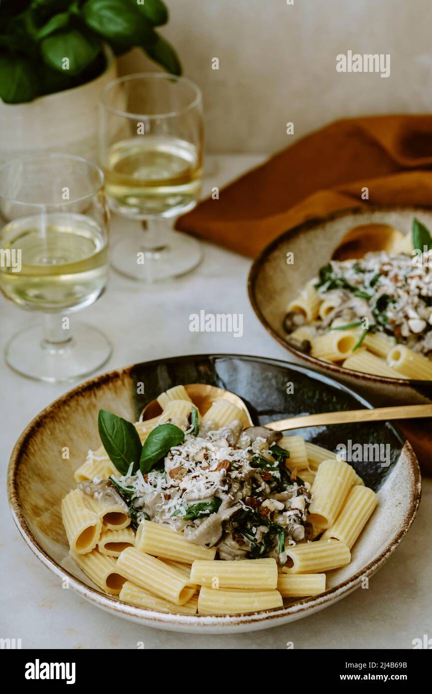 Rigatoni Nudelgericht, mit Creme, Pilz, Spinat, Mandel und geriebenem Parmesan, in handgefertigten Tellern, und Glas Weißwein, auf einem Marmortisch, Basilikum Stockfoto