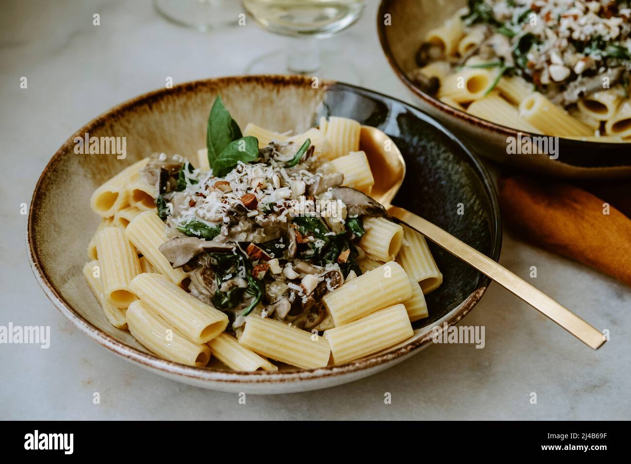 Rigatoni Nudelgericht, mit Creme, Pilz, Spinat, Mandel und geriebenem Parmesan, in handgefertigten Tellern und einem Glas Weißwein, auf einem Marmortisch Stockfoto