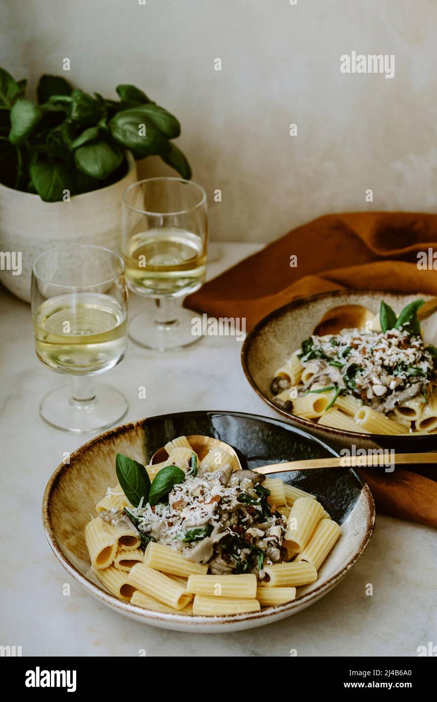 Rigatoni Nudelgericht, mit Creme, Pilz, Spinat, Mandel und geriebenem Parmesan, in handgefertigten Tellern, und Glas Weißwein, auf einem Marmortisch, Basilikum Stockfoto
