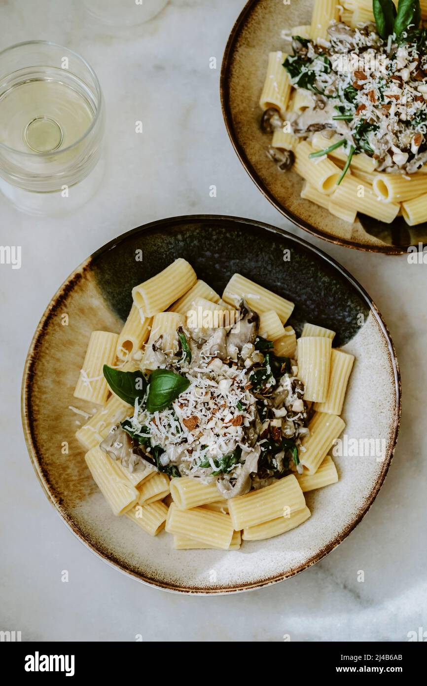 Draufsicht auf zwei Rigatoni-Nudelgerichte, mit Creme, Pilz, Spinat, Mandel und geriebenem Parmesan, in handgefertigten Tellern und einem Glas Weißwein, auf einem Marb Stockfoto