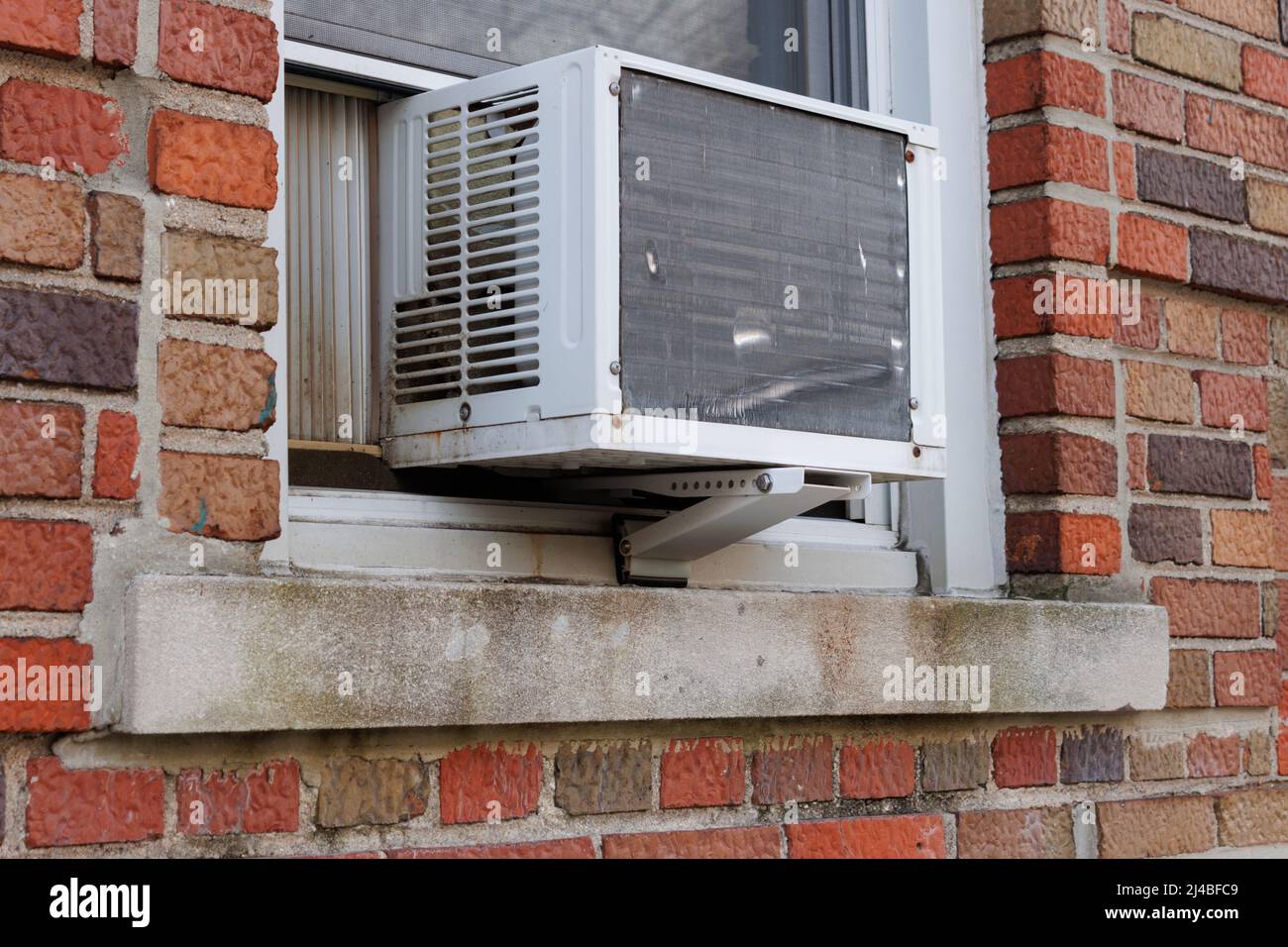 Außenansicht der Klimaanlage, die aus der Fensterbank eines roten Ziegelgebäudes mit einer tragbaren Sicherheitsleiste unter dem Fenster strangiert Stockfoto