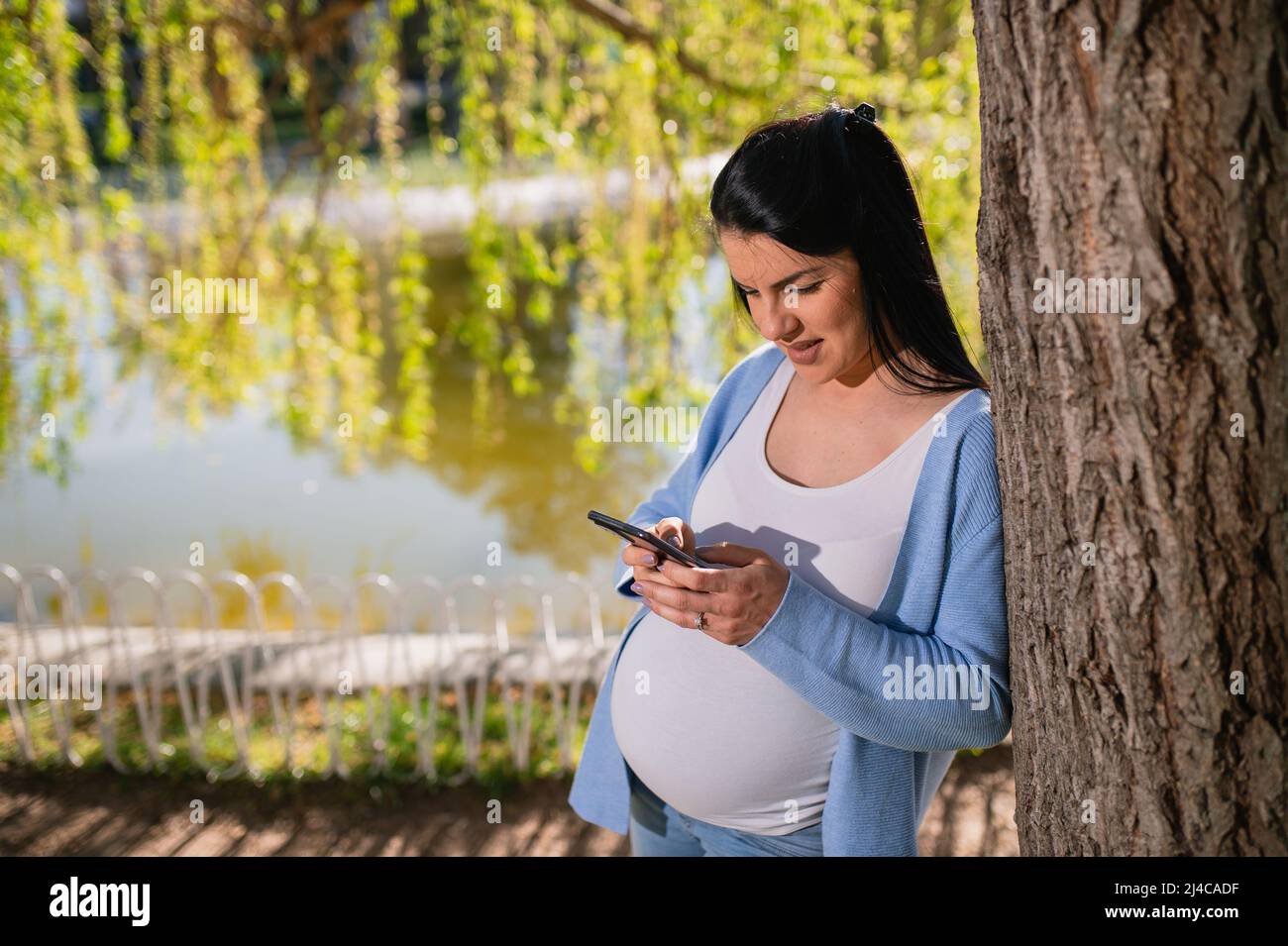 Eine kaukasische Schwangere lehnt sich gut gelaunt an den Baum im Park in der Nähe des Sees. Eine kindgebärende Frau schaut zufrieden auf den Telefonbildschirm. Stockfoto