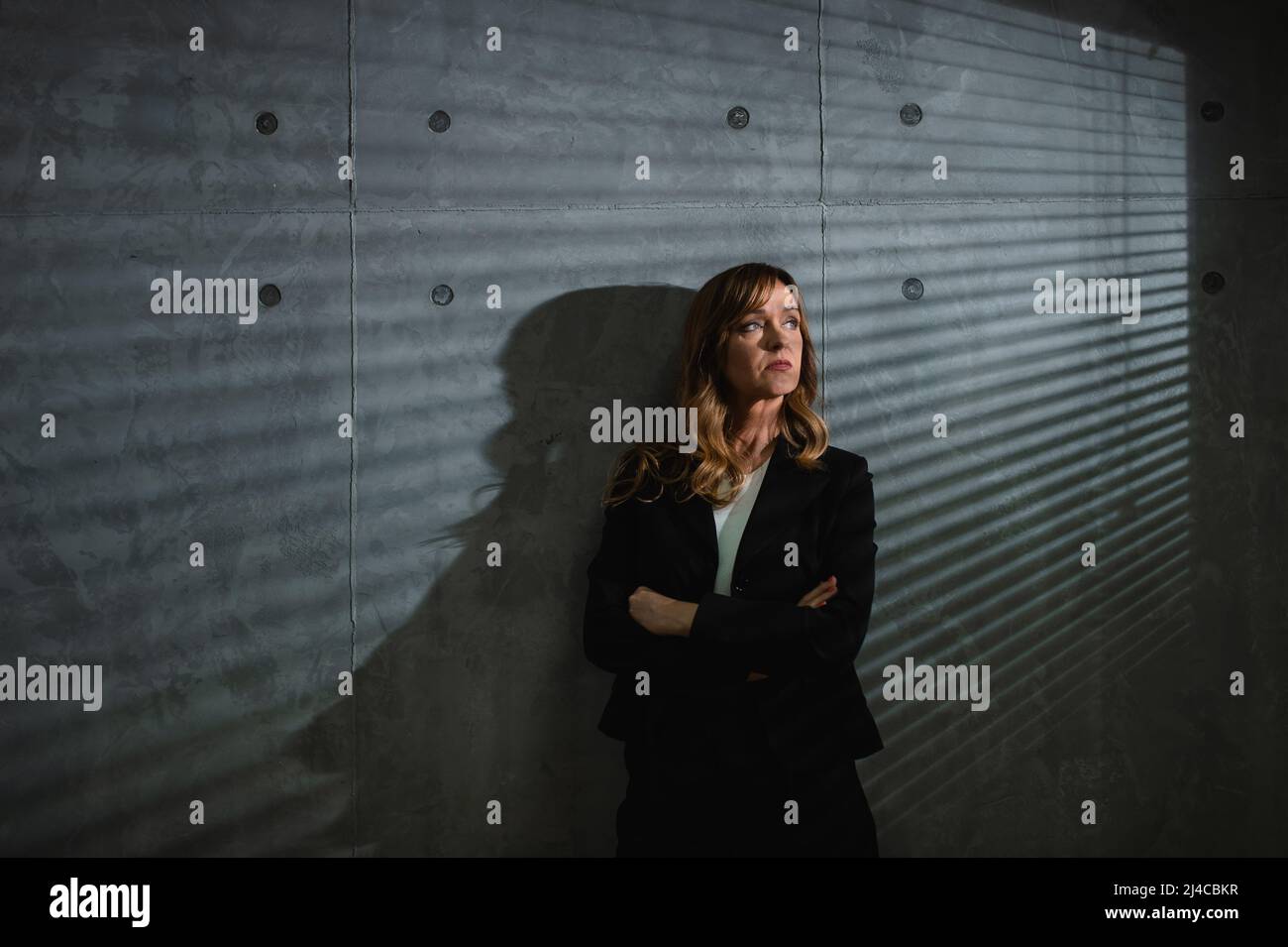 Eine verträumte ältere braunhaarige formell bekleidete kaukasische Frau steht vor grauem Hintergrund in einem abgedunkelten Raum mit gekreuzten Armen. Bllins Shadows. Stockfoto