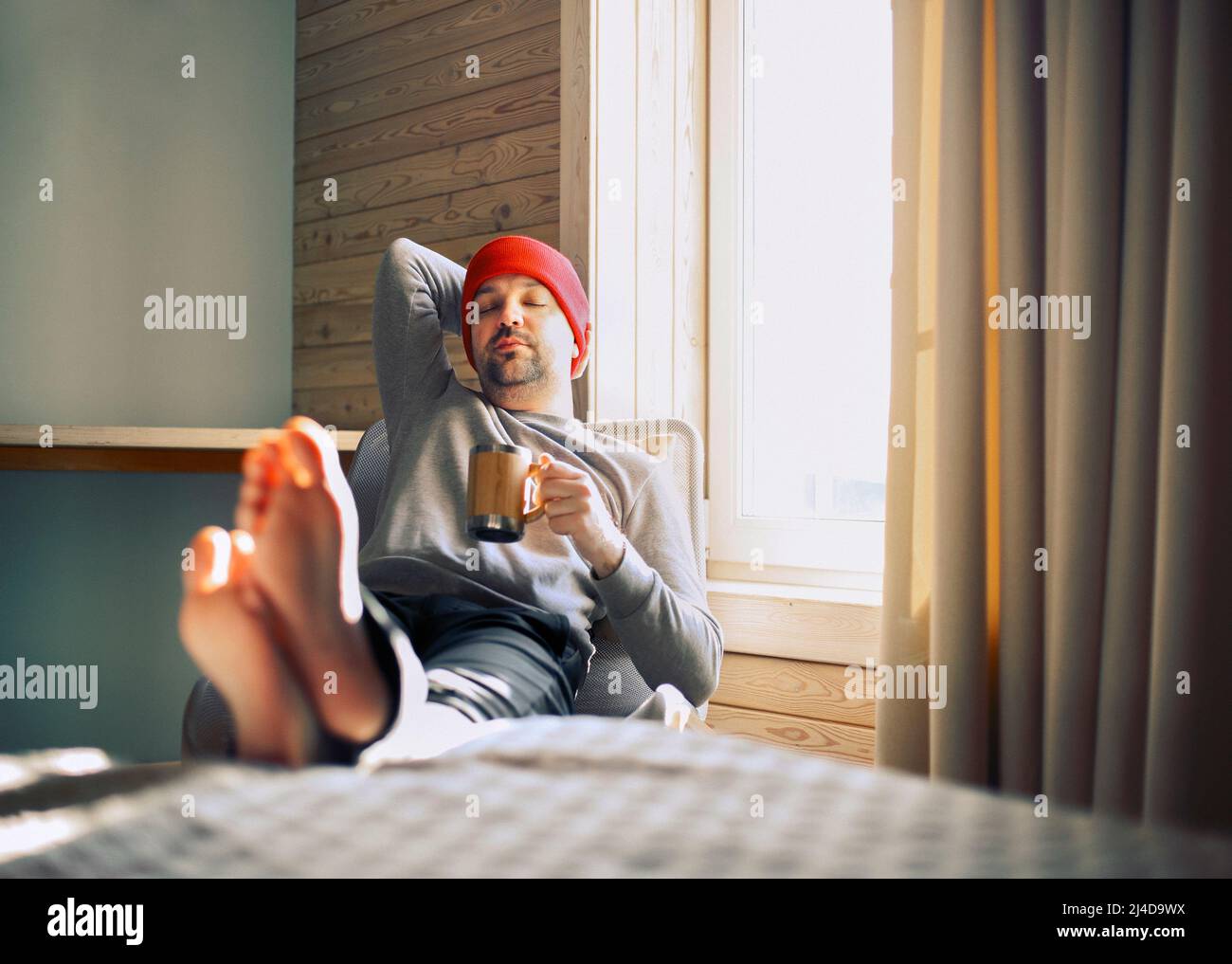 Winter männliche Reisende in roten Hut hält Holzkrug Glühwein oder heißen Tee mit am Fenster in Holz gemütlichen Haus. Mann trinkt Glühwein im sonnigen r Stockfoto