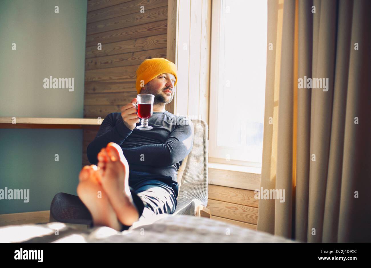 Der männliche Winterreisender in gelbem Hut hält einen Becher Glühwein mit am Fenster in einem gemütlichen Holzhaus. Mann trinkt Glühwein im sonnigen Zimmer Stockfoto