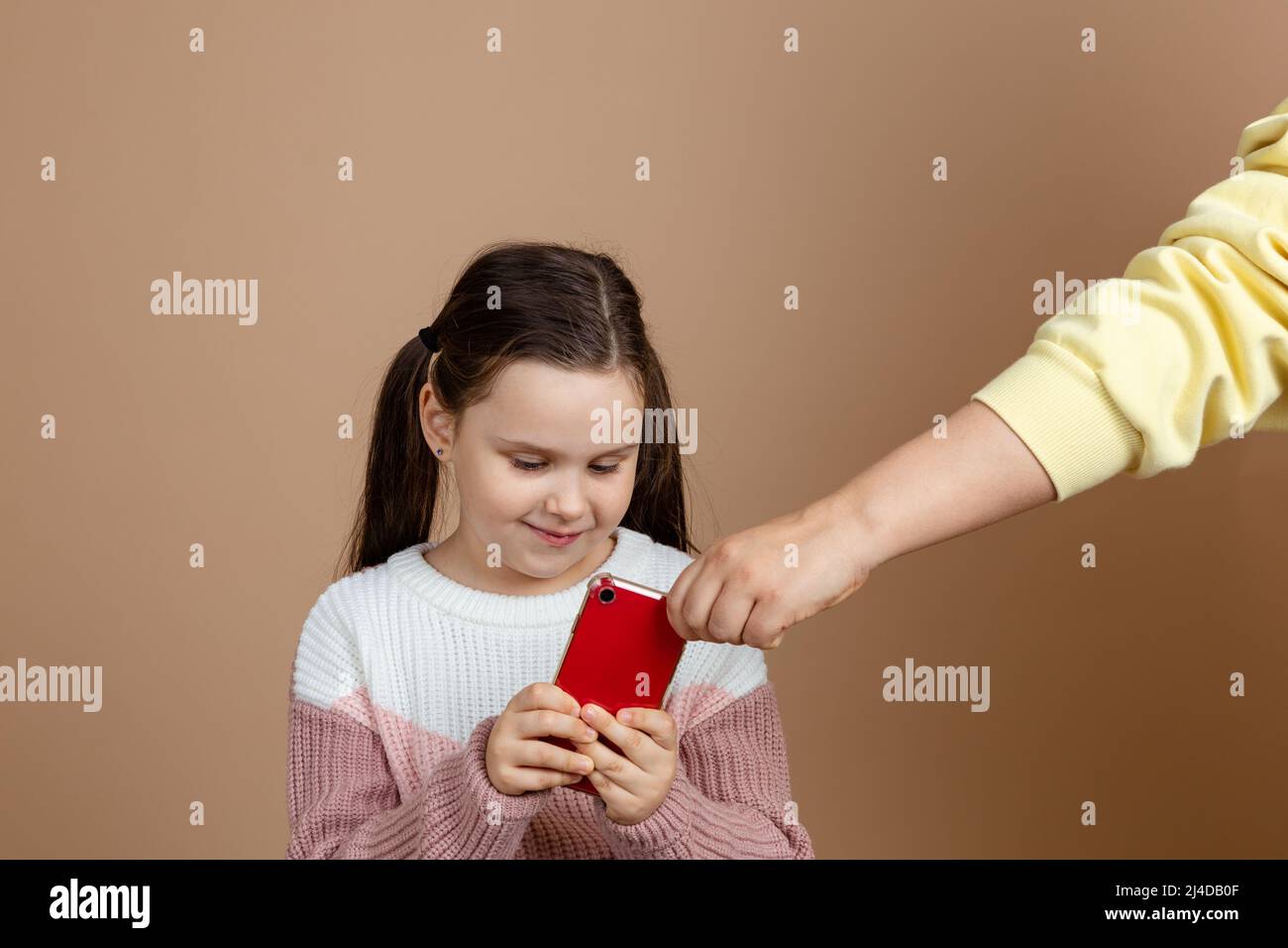 Portrait der Eltern, die das Smartphone aus den Händen der Mädchen nehmen, beige Hintergrund. Lächelnde Tochter hält Telefon und kämpft zurück. Konzept der Prävention von Stockfoto