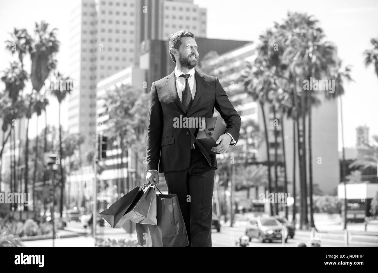 Selbstbewusster Mann in elegantem Anzug mit Einkaufstaschen und geschenkbox draußen spazieren, kaufen Stockfoto