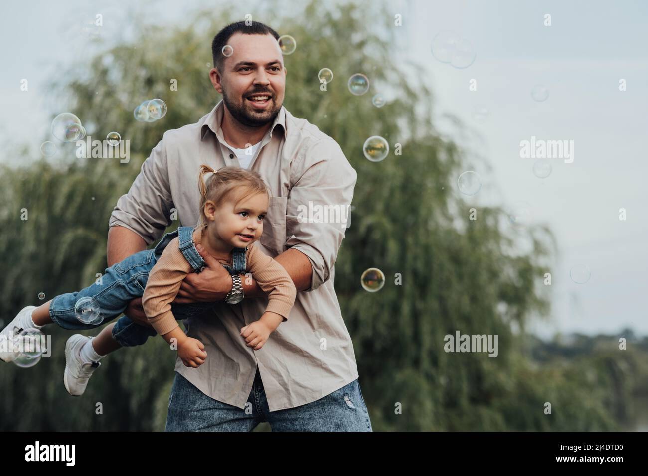 Glücklicher junger Vater hält seine Tochter in den Armen und imitiert Fliegen und spielt mit Blasen im Freien Stockfoto