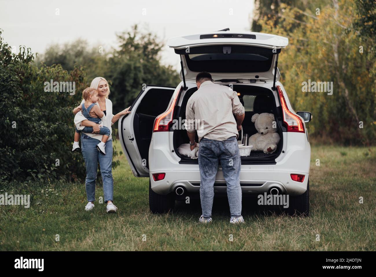 Zwei Eltern mit ihrem kleinen Kind, die sich auf ein Picknick im Freien vorbereiten, junge Familie, die mit dem SUV-Auto unterwegs ist Stockfoto