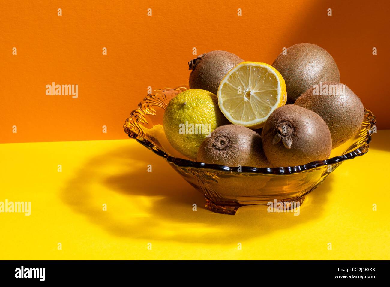 Eine Glasschale mit grünen Kiwi-Früchten, Limette und einer in Scheiben geschnittenen Zitrone auf einem gelben und orangen Hintergrund. Stockfoto