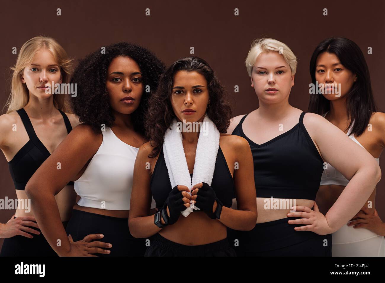 Eine Gruppe von Frauen unterschiedlicher Rasse und Körpergröße in Sportbekleidung, die zusammen stehen. Verschiedene Weibchen betrachten die Kamera vor braunem Hintergrund. Stockfoto