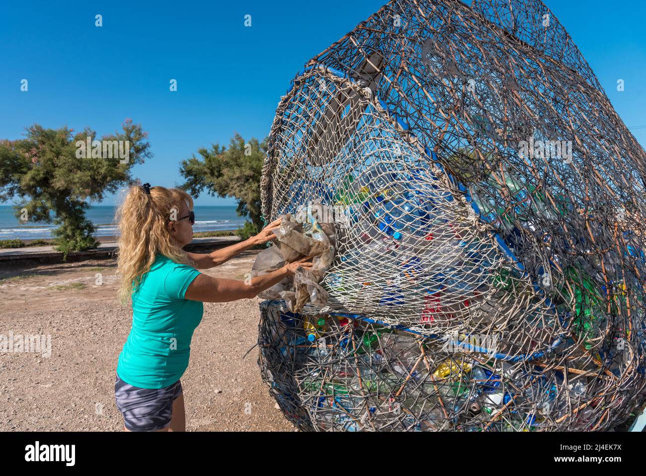 Reife Frau mit Sonnenbrille, die einen Kunststoff in einen sehr großen Abfalleimer voller recycelbarem Material legt. Stockfoto
