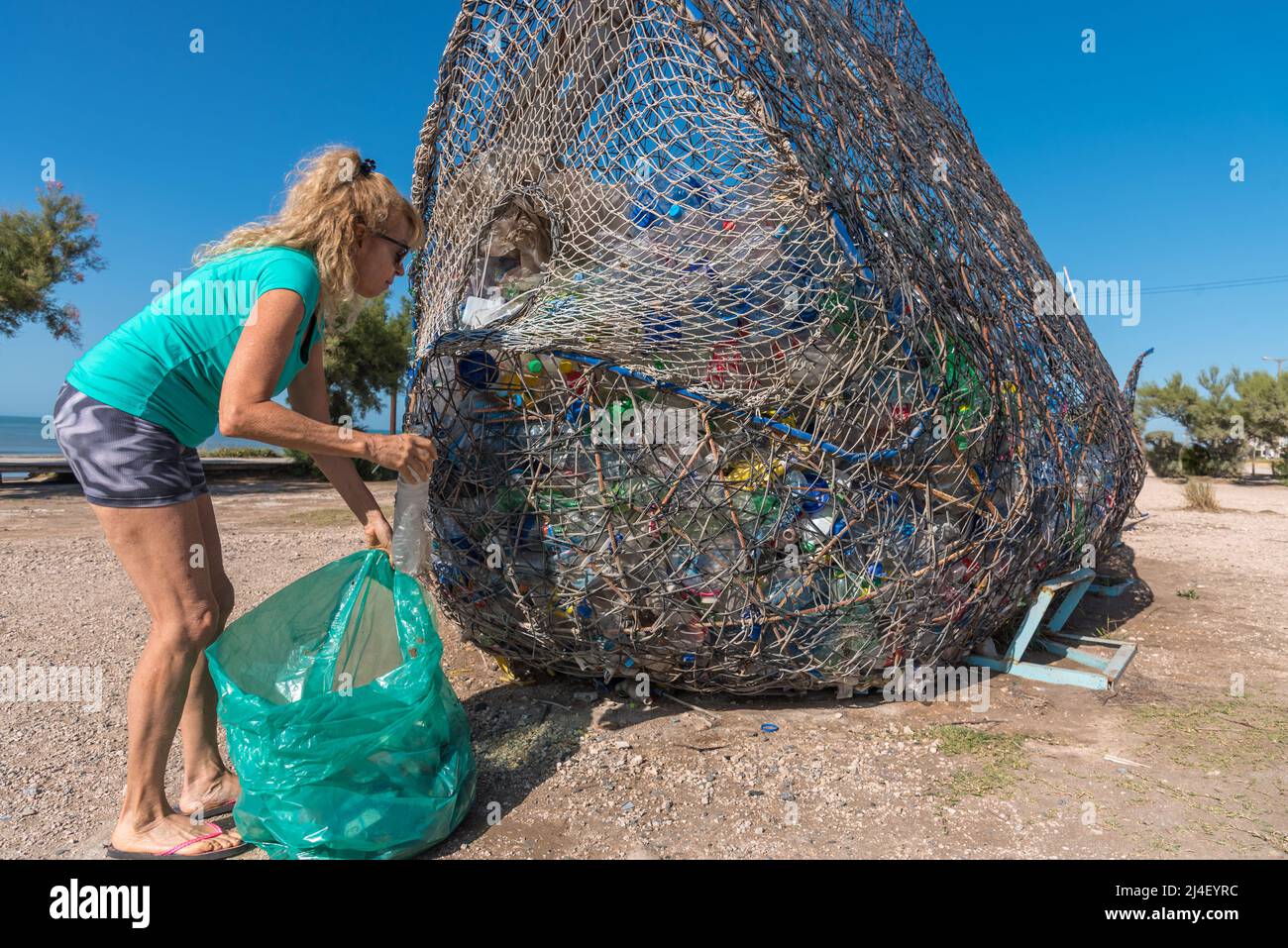 Reife blonde Frau mit Sonnenbrille, die eine Plastikflasche in einen Abfalleimer voller recycelbarem Material legt Stockfoto