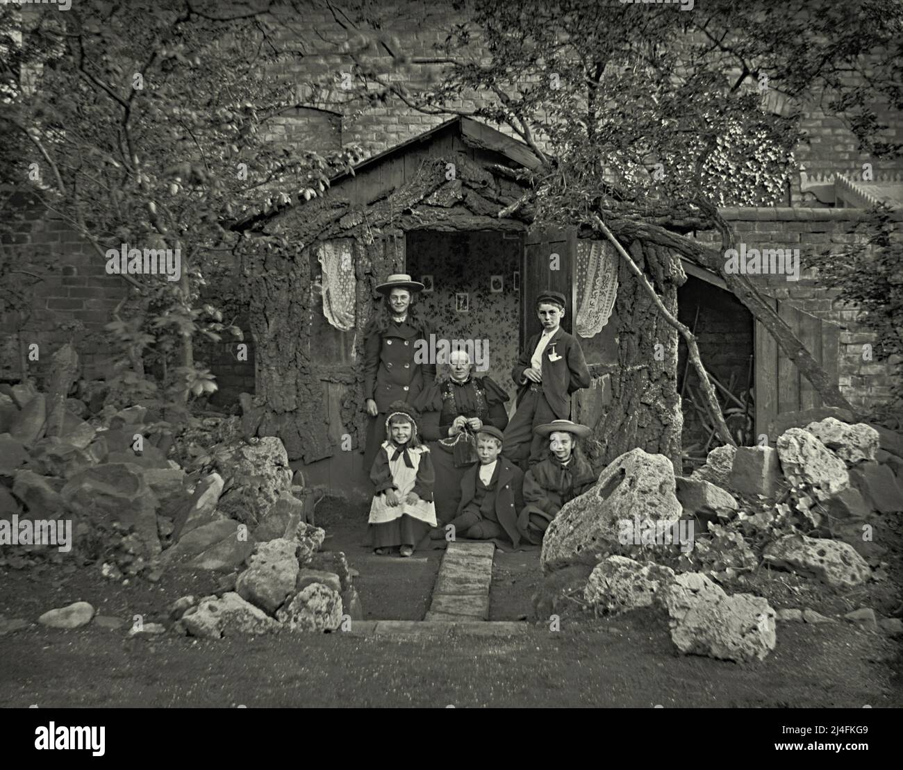 Eine Familiengruppe mit einer Mutter und fünf Kindern posiert vor ihrem viktorianischen Sommerhaus in Großbritannien um 1900. Das rustikale Sommerhaus hat Fenster mit Netzvorhängen, Tapeten und Bilder an der Wand. Daneben befindet sich ein Schuppen mit Stauraum. Vor der Familie befindet sich ein beeindruckender Steingarten mit massiven Felsen und Felsbrocken. Dies ist einem viktorianischen Glasnegativ entnommen – einem Vintage-Foto aus dem Jahr 1800s/1900s. Stockfoto