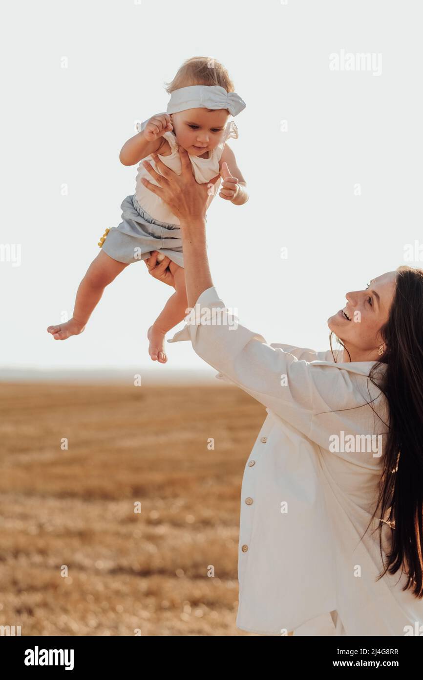 Glückliche Mutter, die ihr Baby in den Armen hält, imitiert die fliegende, junge Mutter, die bei Sonnenuntergang vor der Stadt mit der Tochter eines Kleinkindes spielt Stockfoto