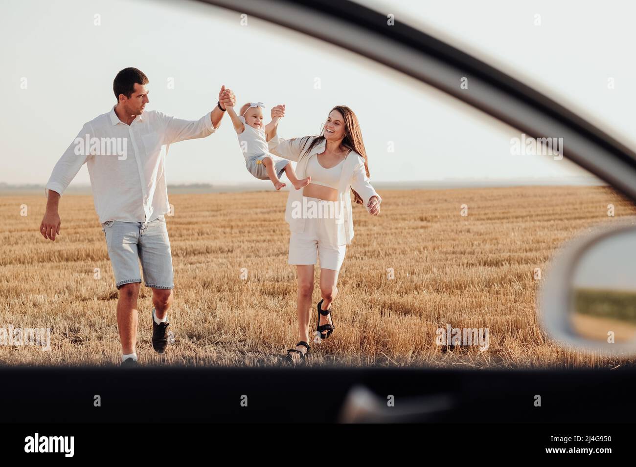 Blick durch das Autofenster, Mama und Papa halten ihre Tochter mit den Händen, während sie bei Sonnenuntergang auf dem Feld spazieren Stockfoto