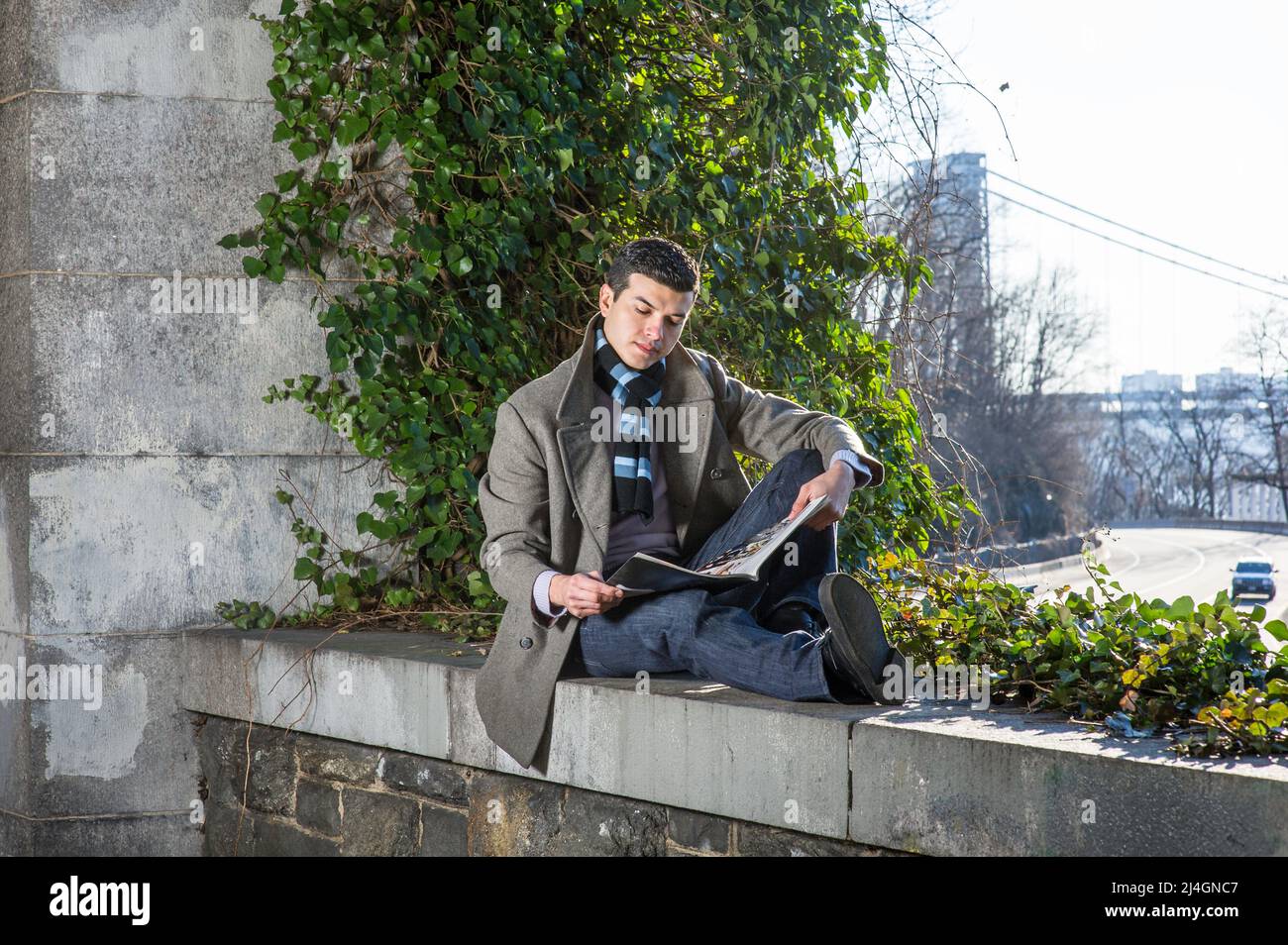 Ein junger Mann, der auf einem großen Rahmen sitzt und vor wilden grünen Pflanzen sitzt, liest ein Magazin Stockfoto